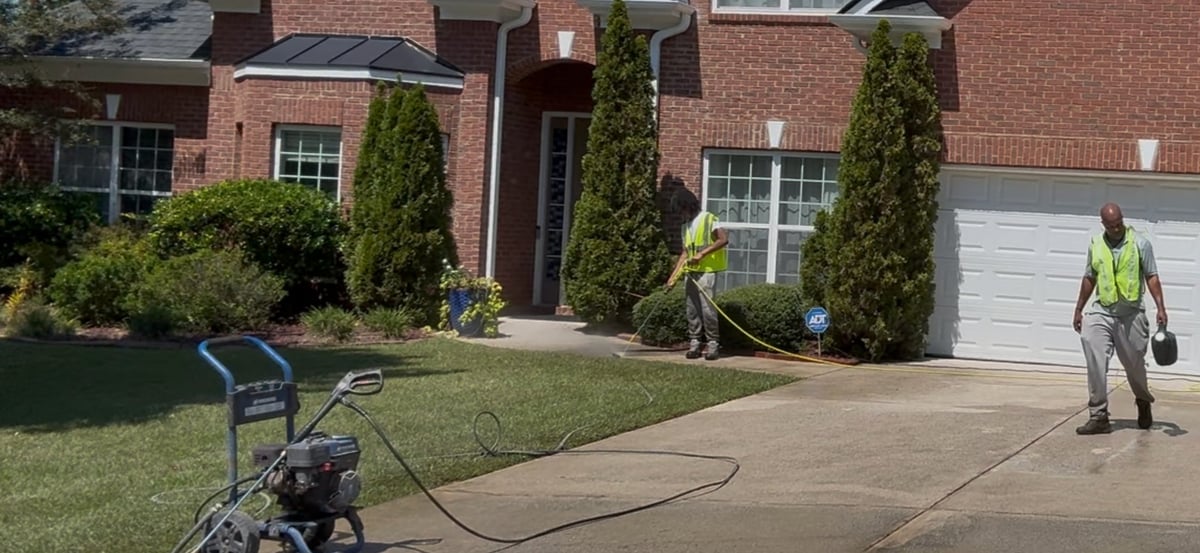 Crew member in safety vest pressure washing a driveway at a brick home with landscaping
