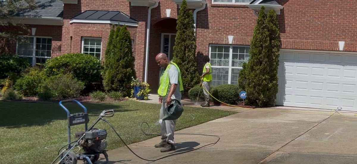 Worker in safety vest power washing a brick house driveway with equipment staged nearby